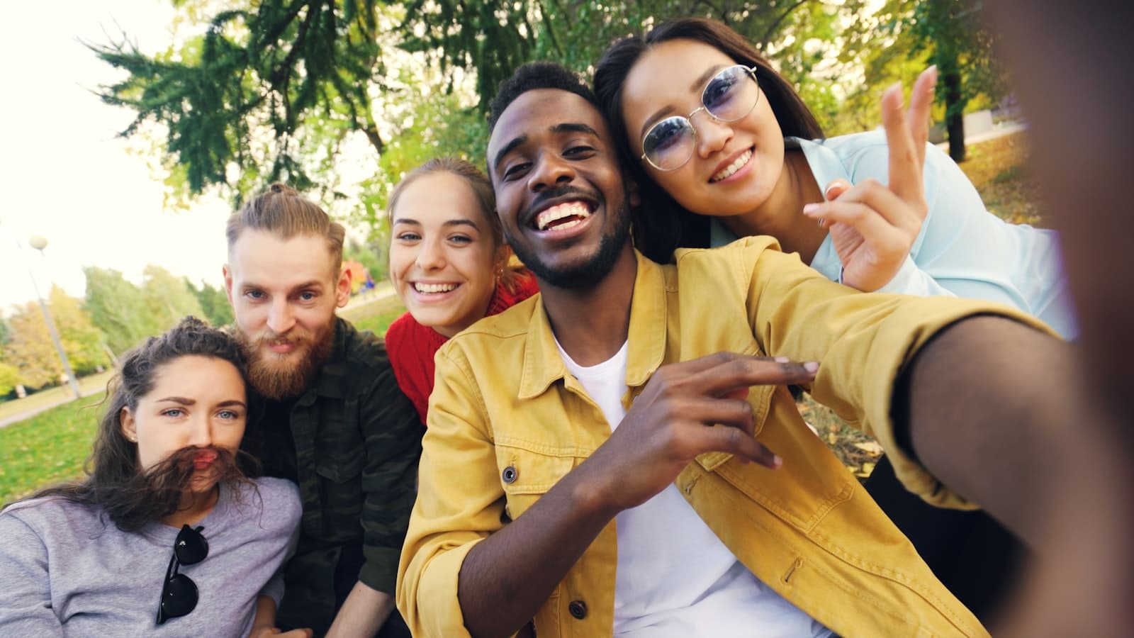 A diverse group of friends smiling together in a group selfie outdoors in a park.